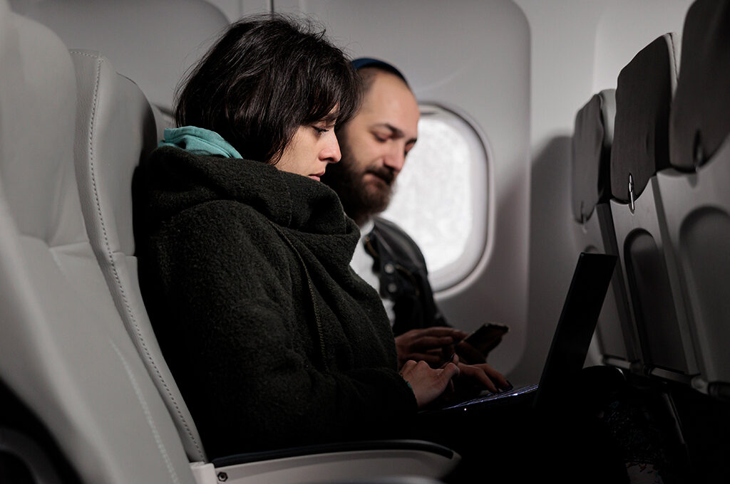 A woman working on her laptop and a man using his smartphone inside an airplane cabin, illustrating productive ideas on what to do on a long haul flight.