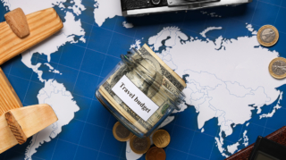 A glass jar labeled "Travel budget" filled with cash, resting on a world map next to a toy wooden airplane and coins, illustrating how to save money for a holiday.