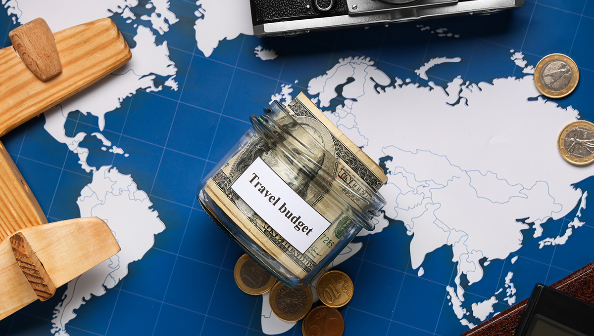 A glass jar labeled "Travel budget" filled with cash, resting on a world map next to a toy wooden airplane and coins, illustrating how to save money for a holiday.