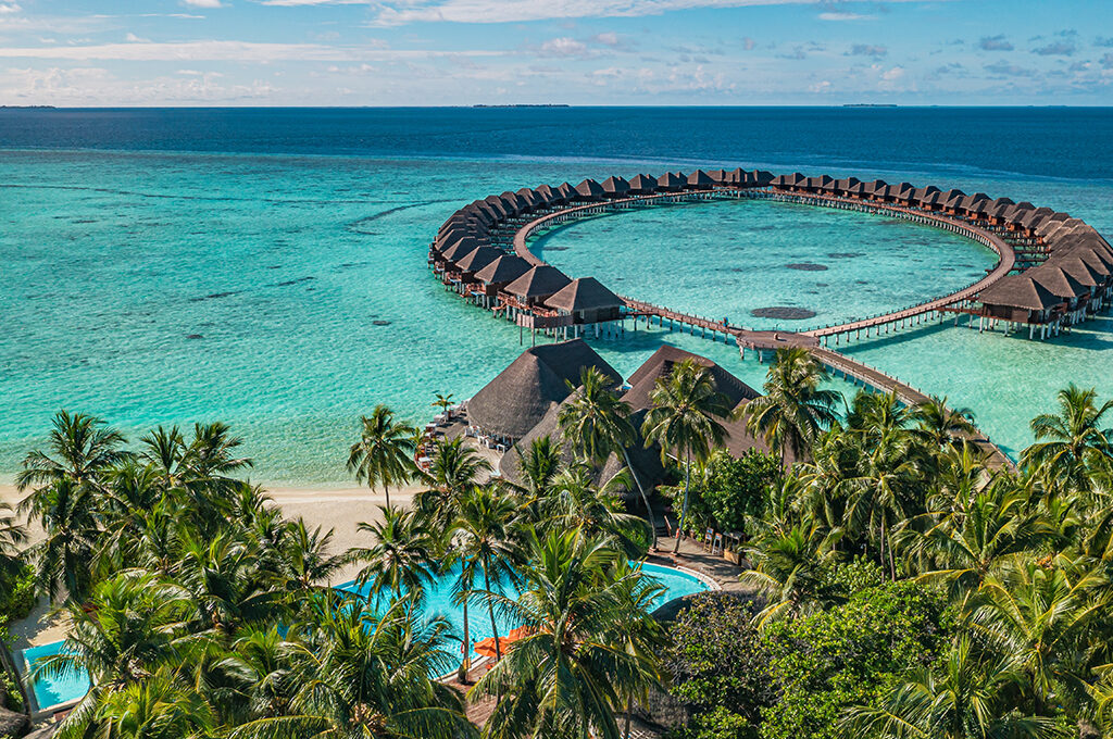 Aerial view of an overwater bungalow resort arranged in a circular shape over turquoise water, next to a tropical beach with palm trees and a swimming pool, representing one of the most popular holiday destinations.