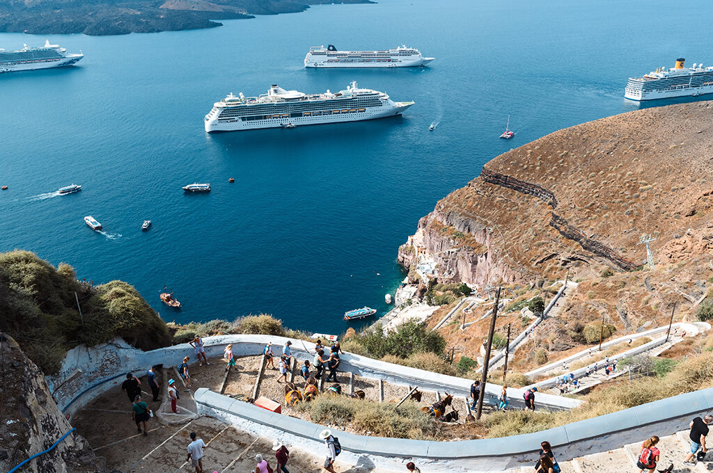 High-angle view of multiple large cruise ships anchored in a deep blue sea near steep volcanic cliffs and a scenic winding pathway stairs, characteristic of the most popular holiday destinations.