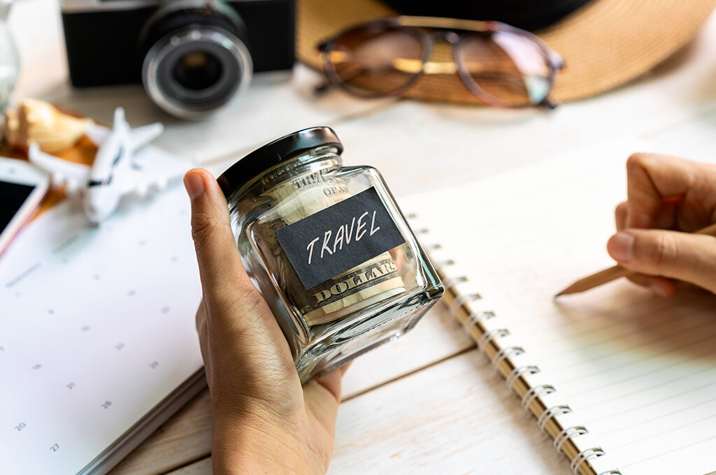 A person holding a jar filled with dollars labeled "TRAVEL" while writing notes in a planner, demonstrating steps on how to save money for a holiday.