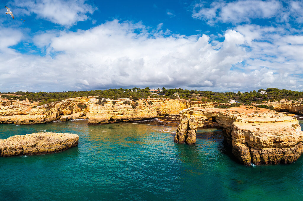 Scenic view of golden coastal rock formations and arches meeting clear turquoise waters under a cloudy blue sky in Algarve is one of the most popular holiday destinations.