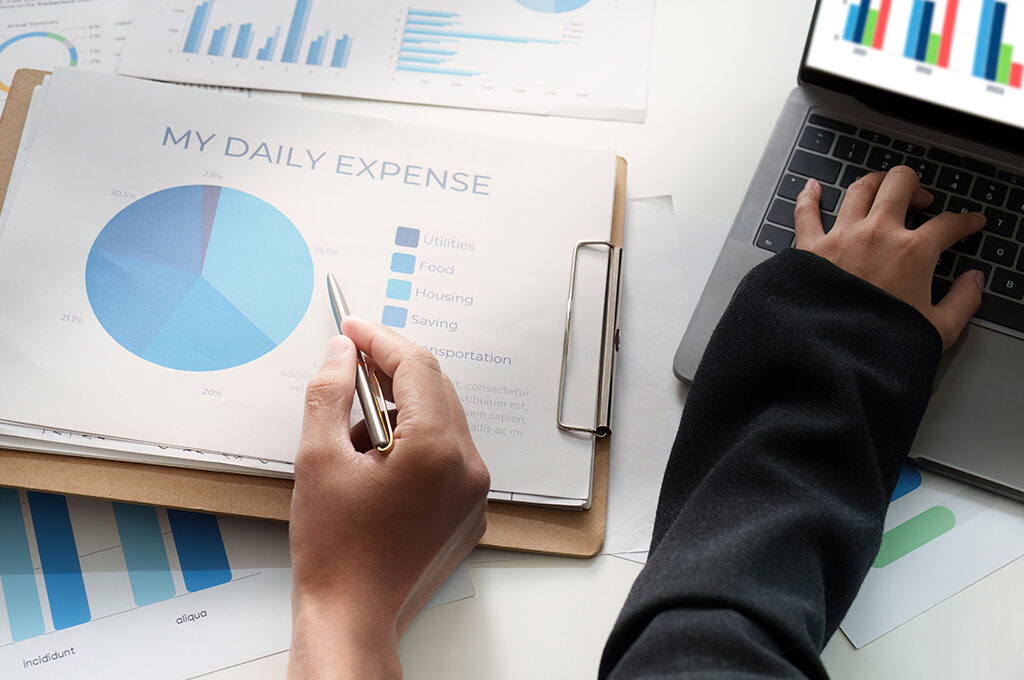 A person working on a laptop and analyzing a pie chart labeled "MY DAILY EXPENSE" to help figure out how to save money for a holiday.
