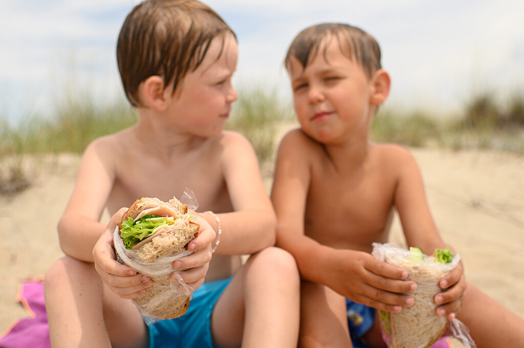 Two young children sitting on a beach holding sandwiches wrapped in plastic, representing budget-friendly travel choices.