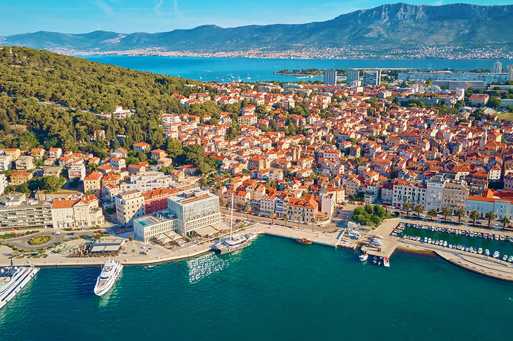Aerial view of a historic coastal town with red-tiled roofs, a marina with docked boats, and mountains in the background as one of the most popular holiday destinations.