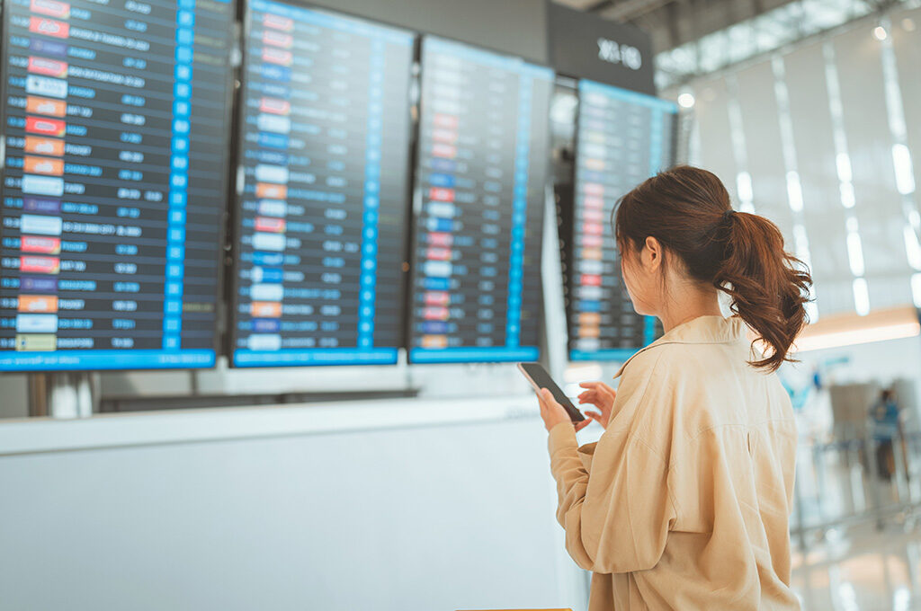 Woman checking her smartphone in front of a blue airport flight information departure board.