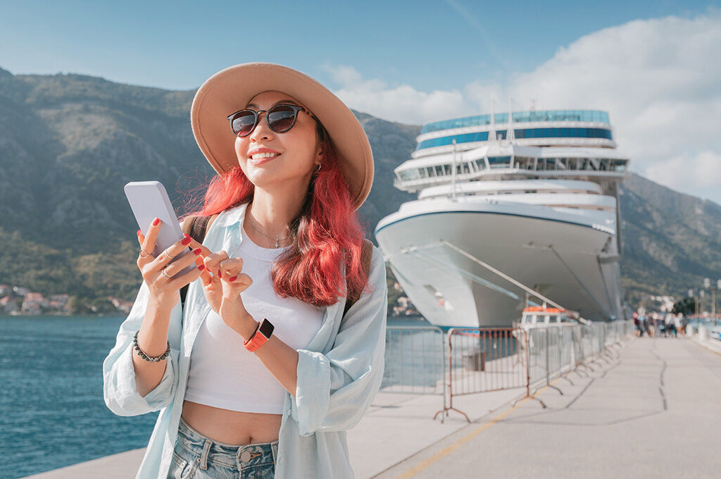 A traveler checking a smartphone on a pier with a cruise ship docked in the background, highlighting trip planning when researching when is the best time to book a cruise.