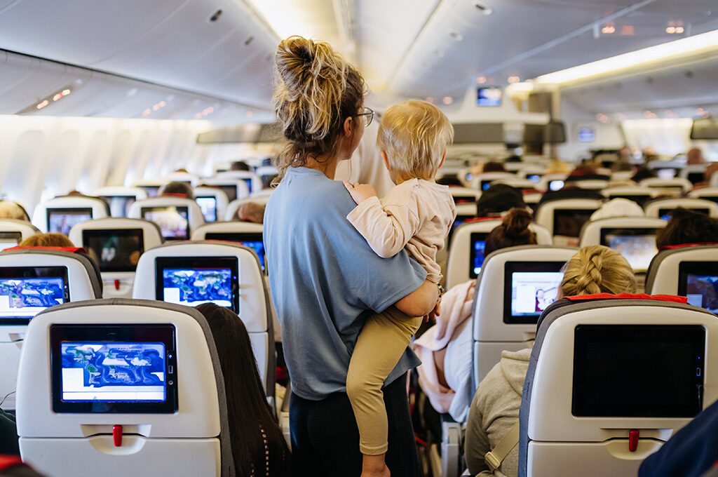 Mother carrying her toddler down the aisle of a crowded airplane economy cabin.