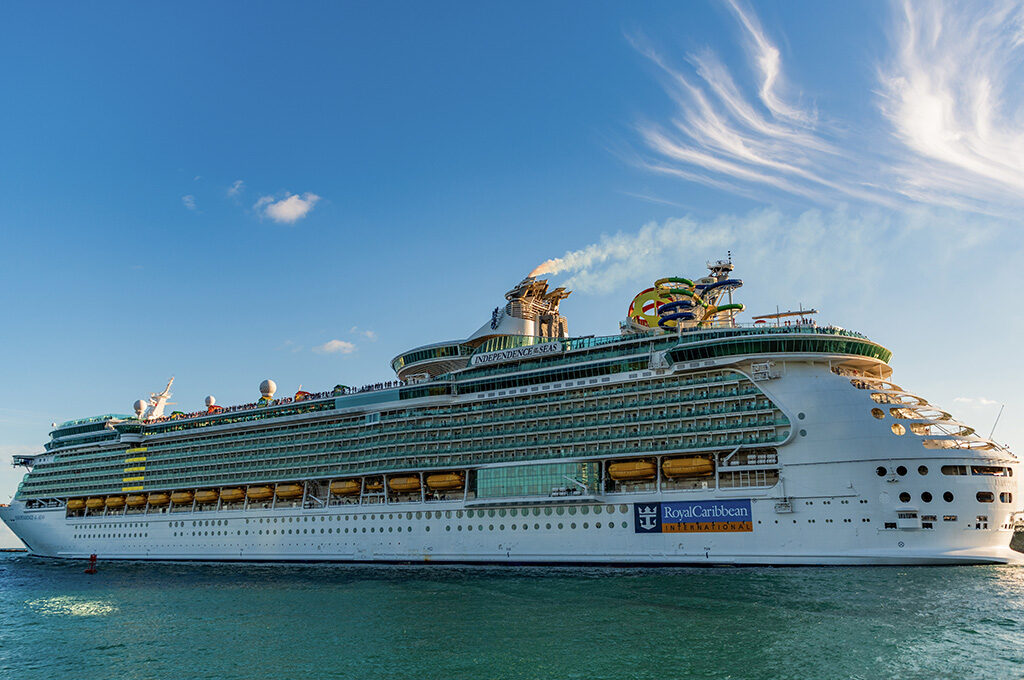 A large white cruise ship with waterslides on the top deck cruising on blue water under a clear sky.
