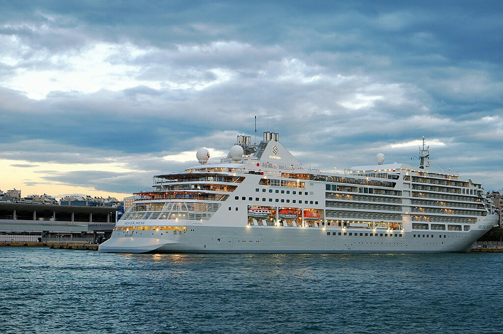 A luxury Silversea cruise ship anchored near a port city under a cloudy sunset sky, showcasing options included in the best cruise lines from Southampton.