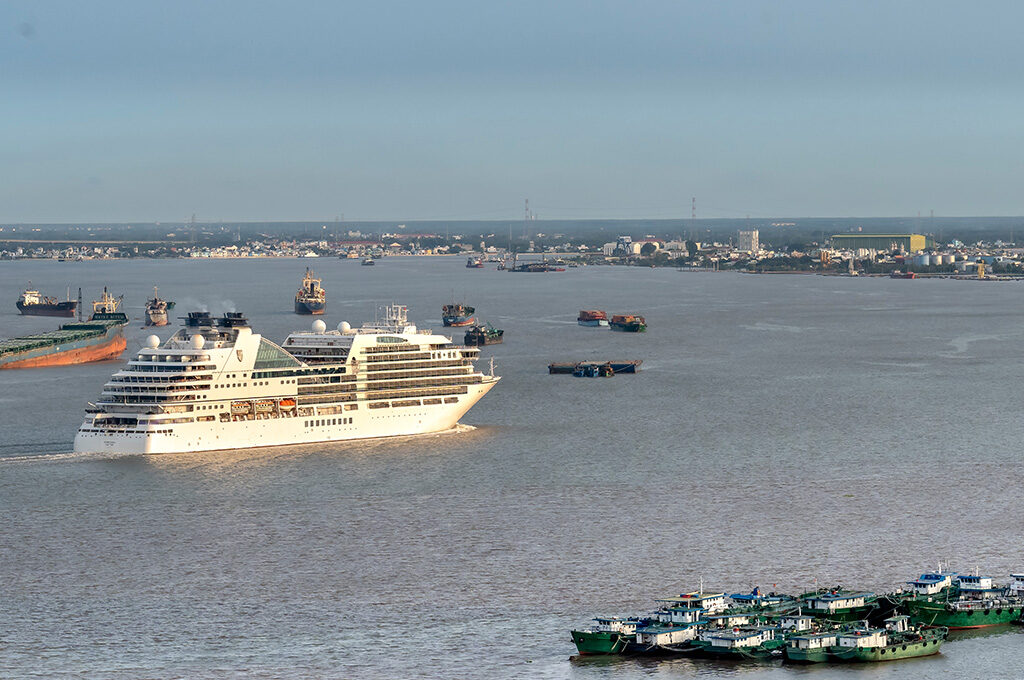An aerial view of a luxury cruise ship navigating a busy river channel filled with commercial cargo vessels, representing routes connected to the best cruise lines from Southampton.