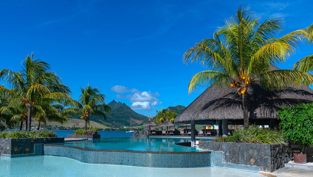 Infinity pool surrounded by palm trees and thatched-roof pavilions with mountain views at one of the best hotels in mauritius.