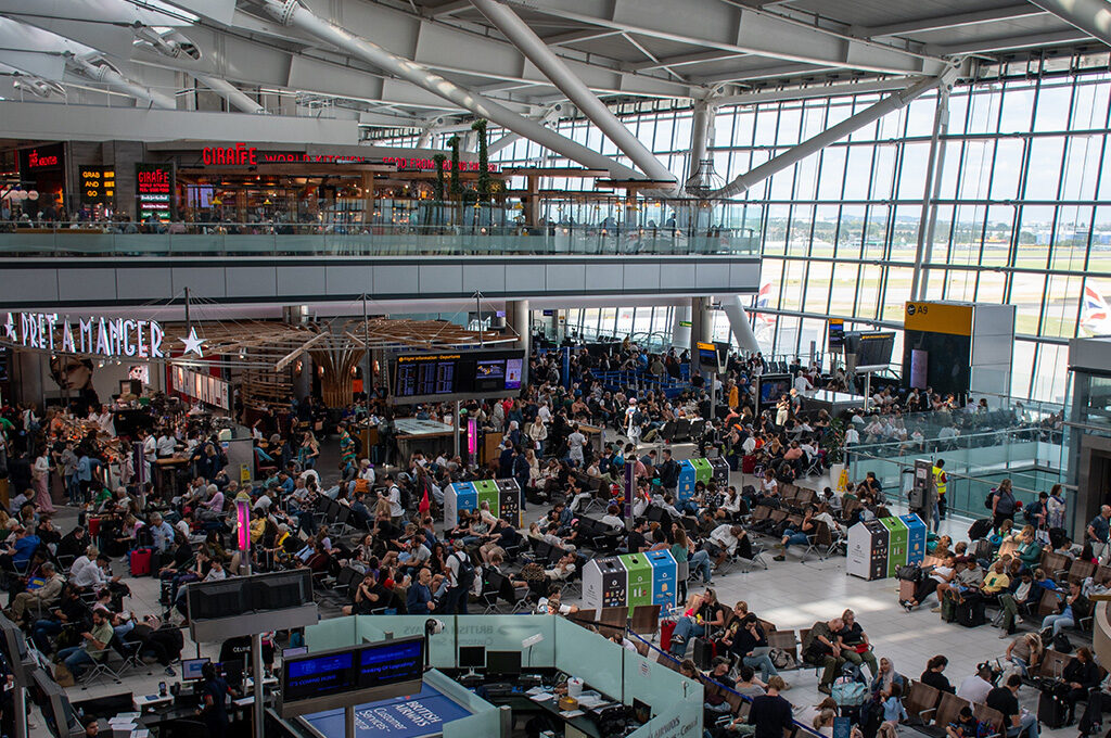 Overview of the crowded departure lounge at London Heathrow Terminal, one of the busiest airports in Europe, featuring retail shops like Harrods, dining options, and seating areas under a arched glass ceiling.